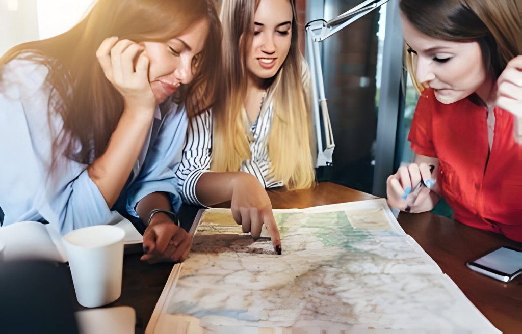 Three girlfriends planning their vacation sitting at table around map choosing the destination
